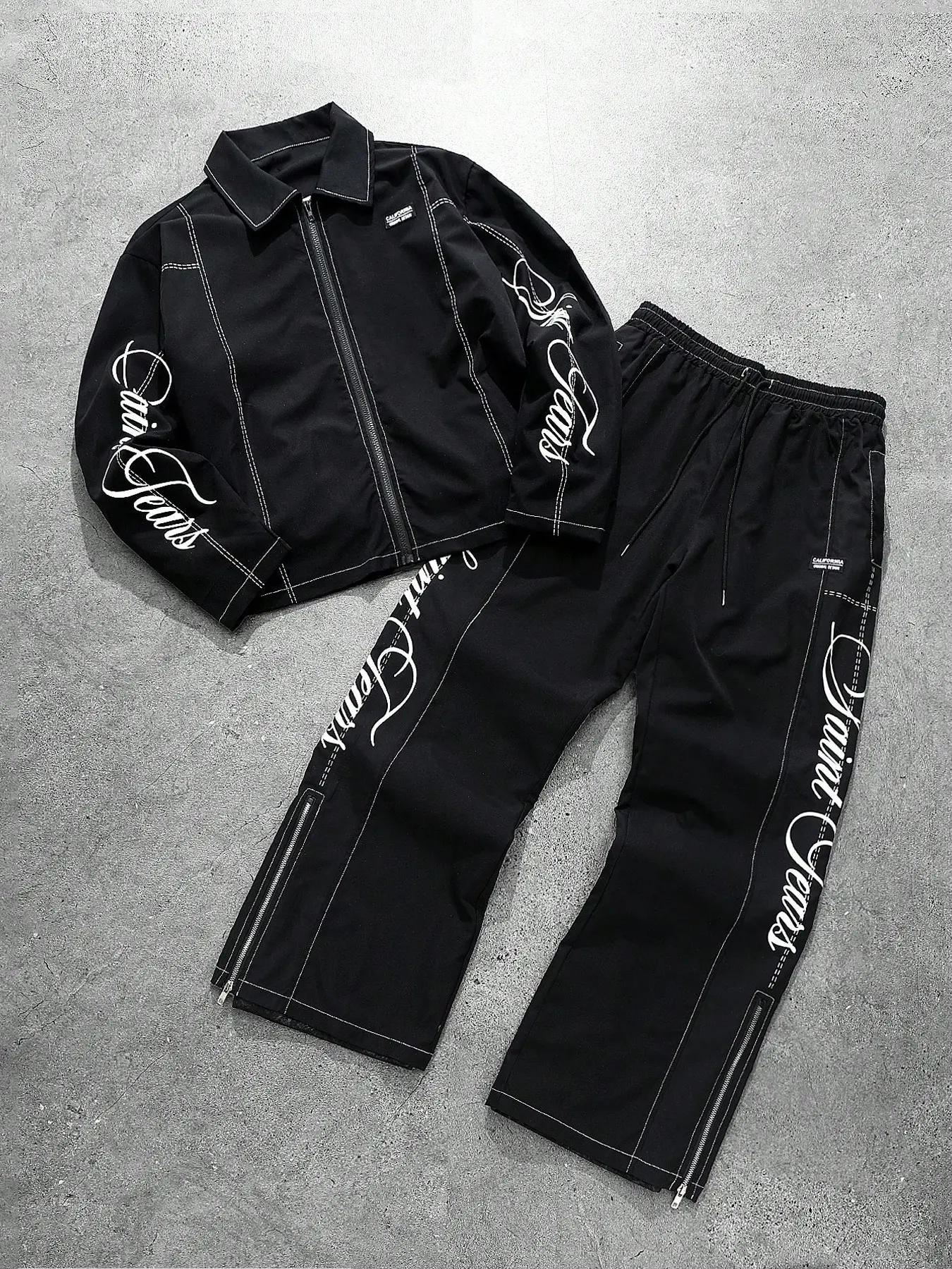This two-piece men's tracksuit features a zip-up jacket with visible stitching and graphic lettering, paired with drawstring flared trousers. A millennial streetwear style set, suitable for skateboarding and everyday casual wear.