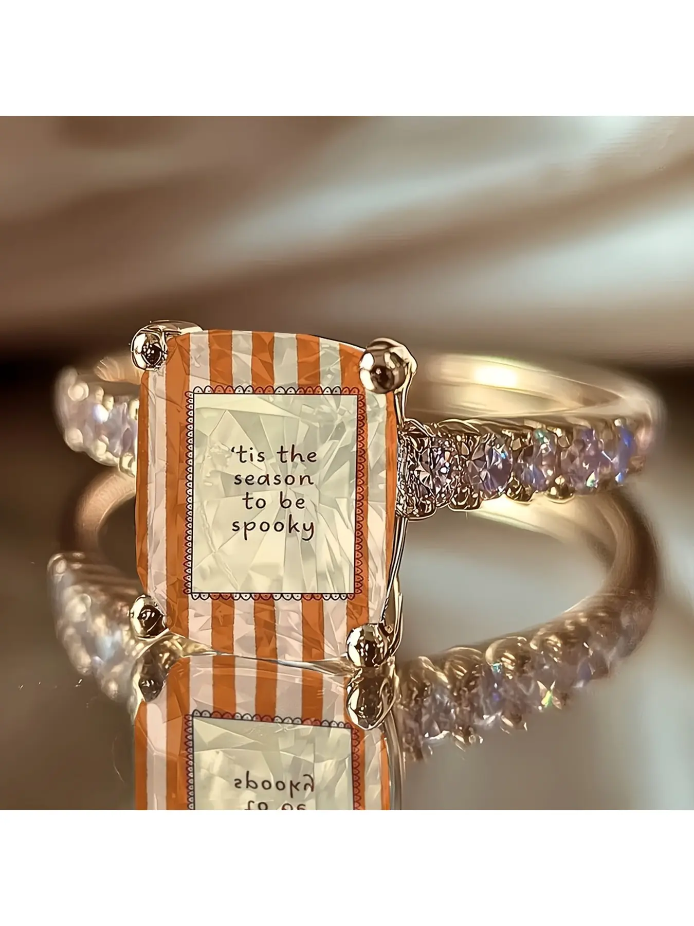 Orange and white vertical striped Halloween text printed ring, Costume rings. The middle square frame is printed with the slogan 'It's the season to be spooky'. The transparent cut surface base, paired with a gold-studded golden ring arm,